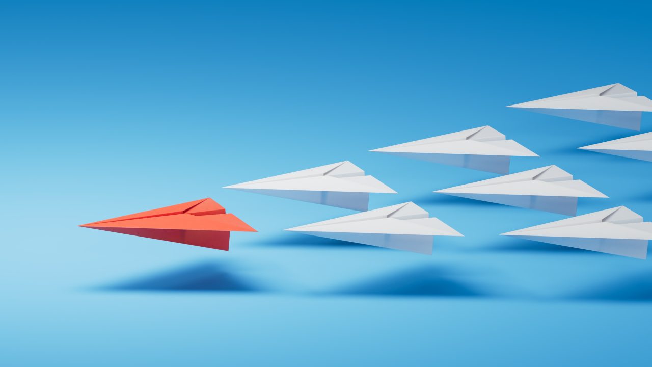 A red paper plane leading 7 white paper planes.