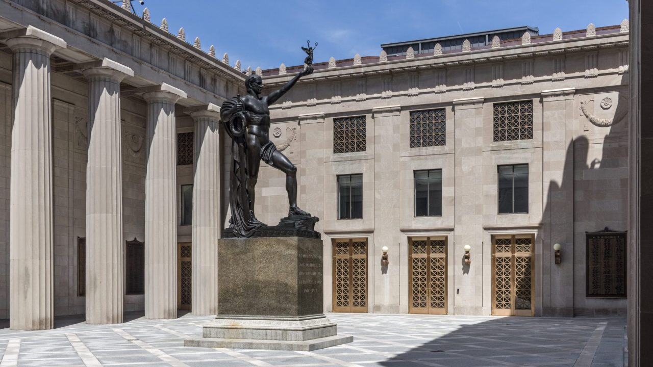 Exterior courtyard of a neoclassical building featuring tall stone columns, ornate gold entrance doors and a large bronze statue on a pedestal under a clear blue sky.