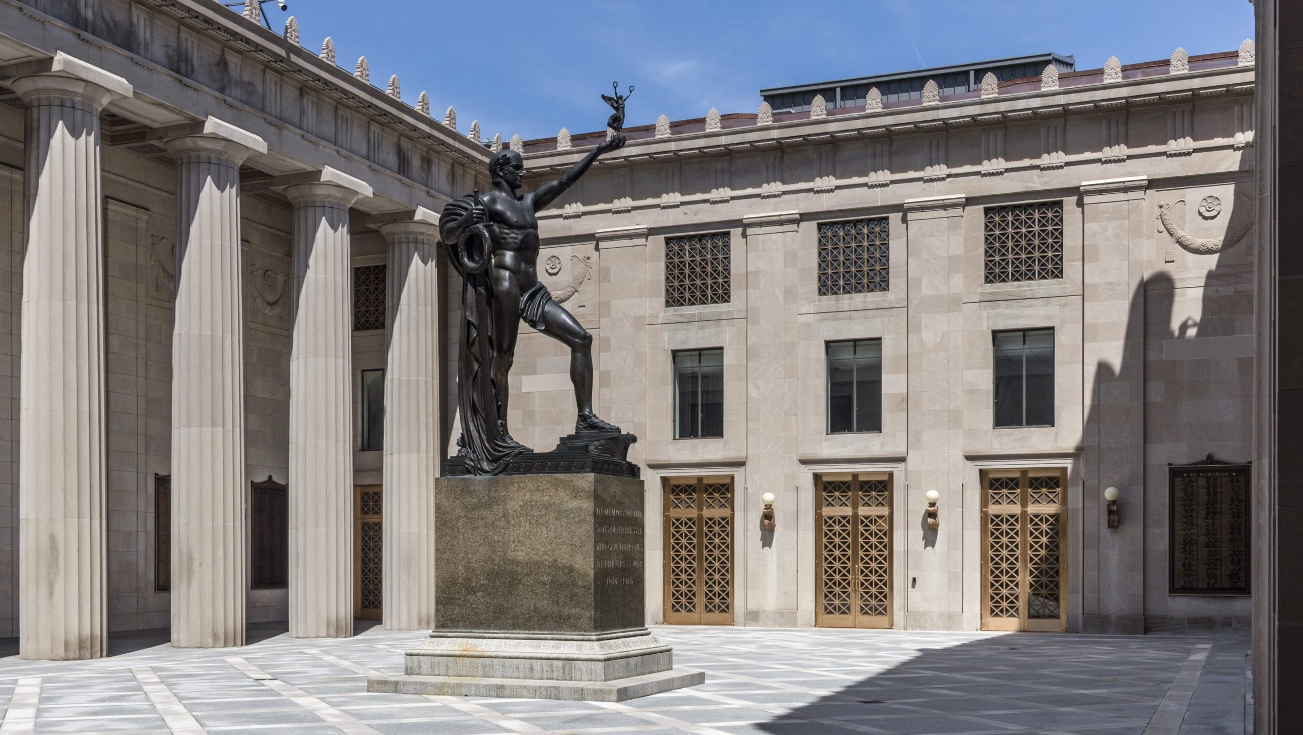 Exterior courtyard of a neoclassical building featuring tall stone columns, ornate gold entrance doors and a large bronze statue on a pedestal under a clear blue sky.
