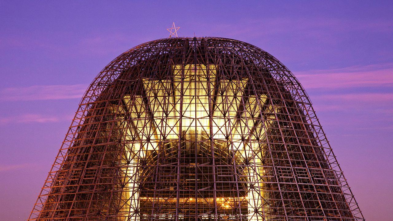 Hangar One at NASA's Ames Research Center illuminated at dusk, showcasing its massive steel lattice frame structure topped with a star against a purple twilight sky.