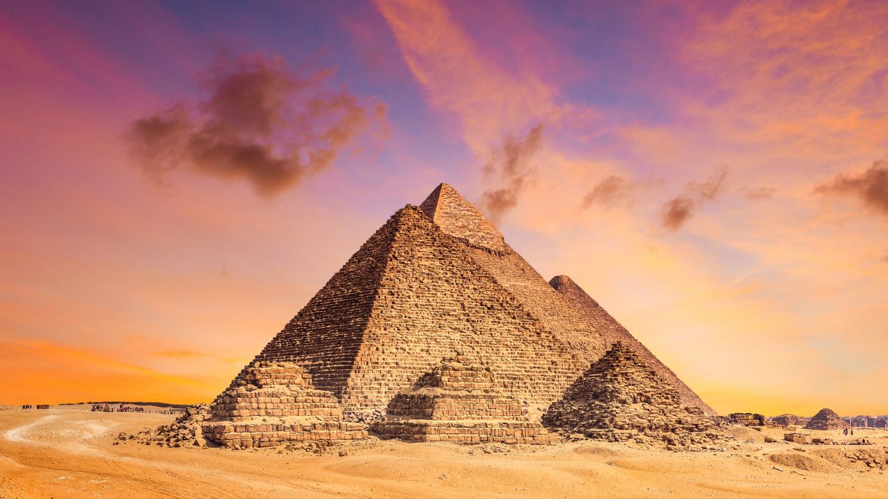 The Pyramids of Giza rising from the Egyptian desert under a vivid orange and purple sunset sky.