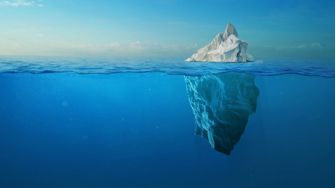 An iceberg floating in open ocean, showing both its small visible peak above the waterline and its much larger mass submerged below the surface.