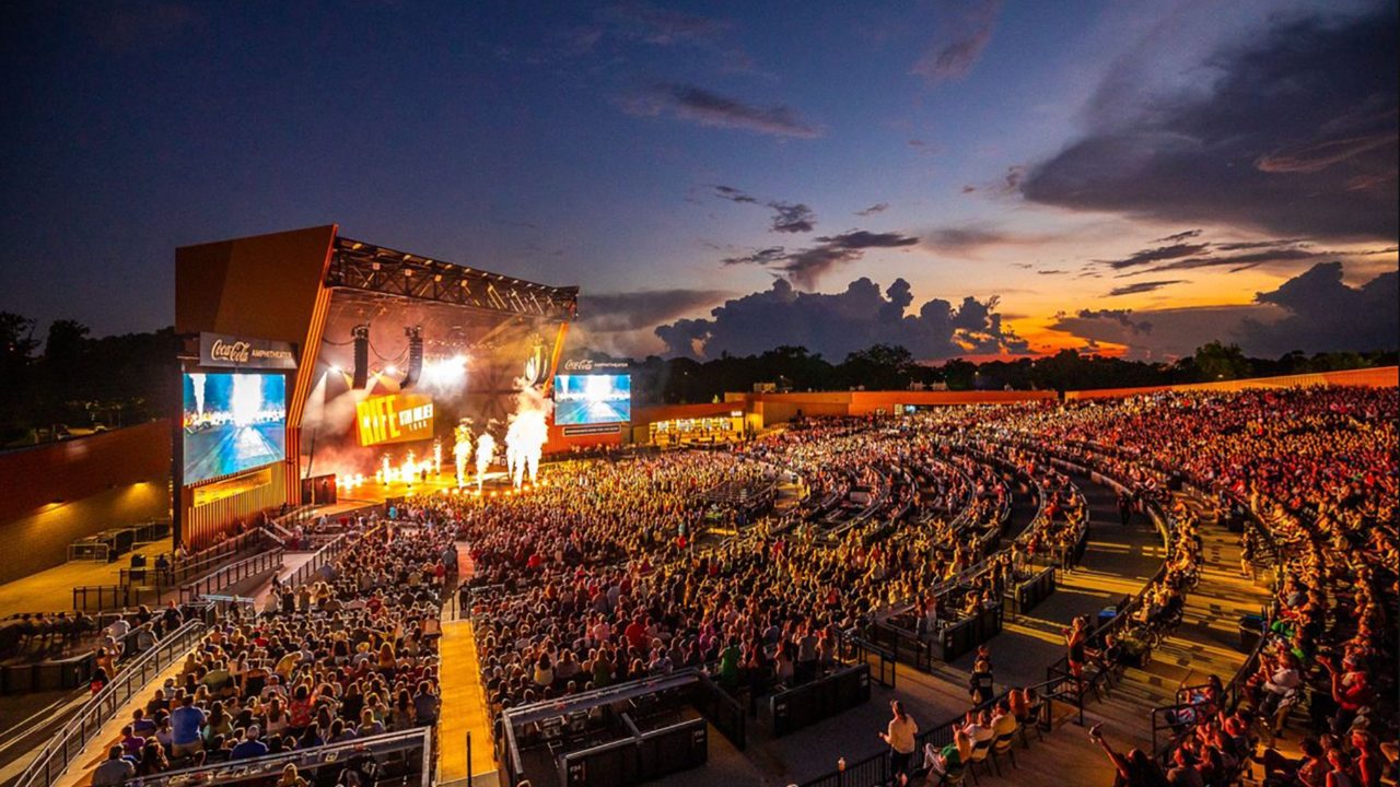 A packed crowd at the Coca-Cola Amphitheater during a live concert at dusk, with a brightly lit stage, large video screens and pyrotechnics against a dramatic sunset sky.