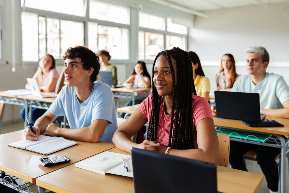 A diverse group of students sitting at desks in a bright classroom, attentively listening with notebooks and laptops open in front of them.