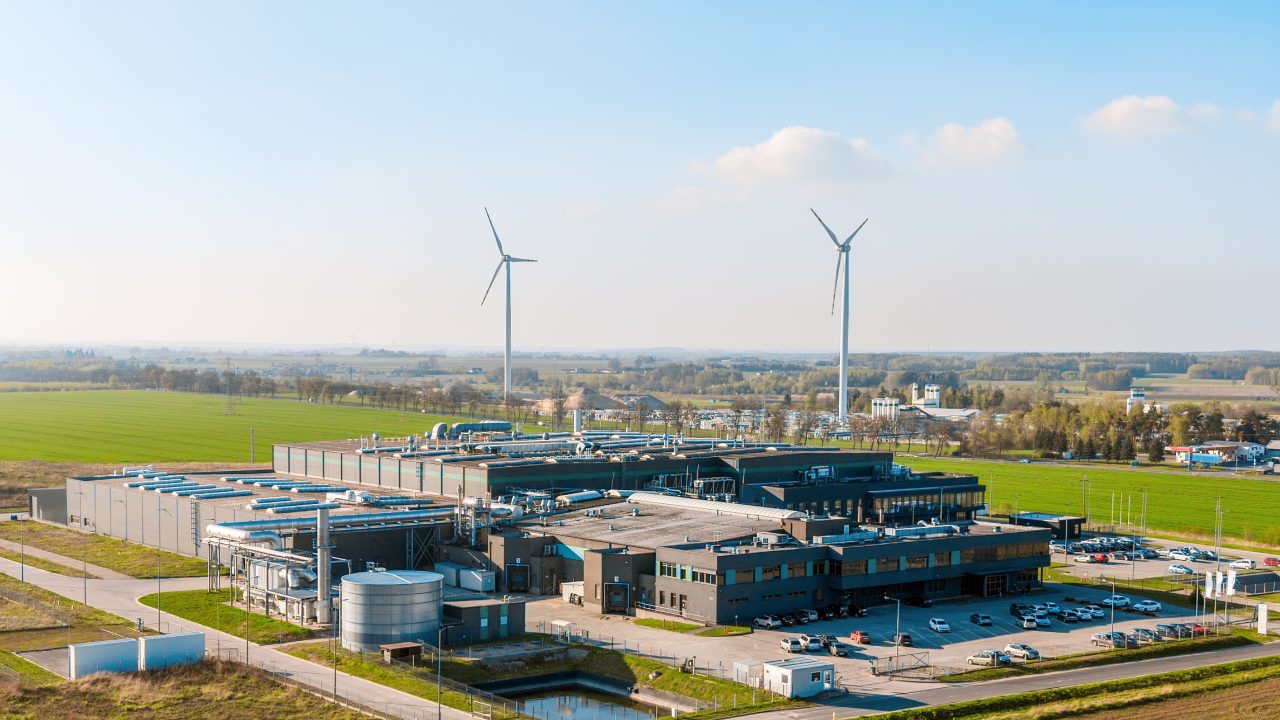 Aerial view of a large industrial facility with rooftop mechanical systems and a storage tank, surrounded by green fields and two wind turbines under a clear blue sky.