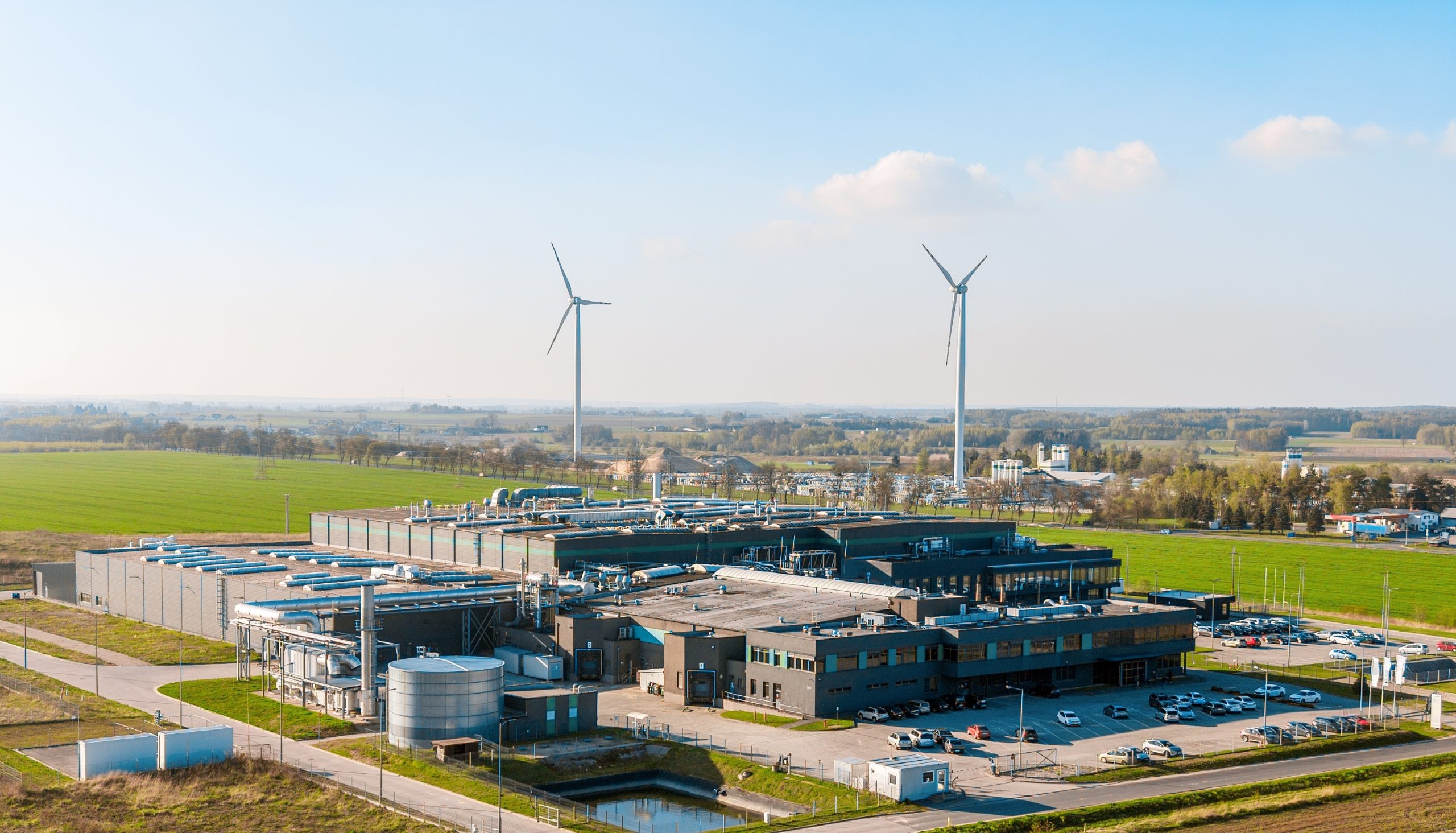 Aerial view of a large industrial facility with rooftop mechanical systems and a storage tank, surrounded by green fields and two wind turbines under a clear blue sky.