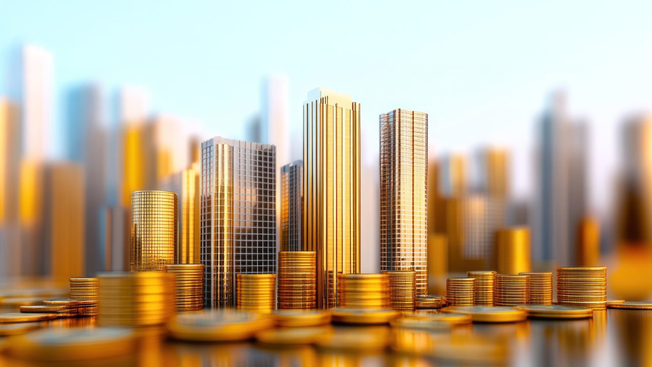 A conceptual image of golden skyscrapers rising among stacks of gold coins, with a blurred city skyline in the background and reflections on a glossy surface.