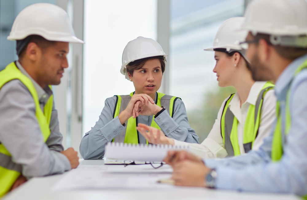 A diverse group of four construction professionals in hard hats and high-visibility vests in discussion around a table with notebooks and blueprints.