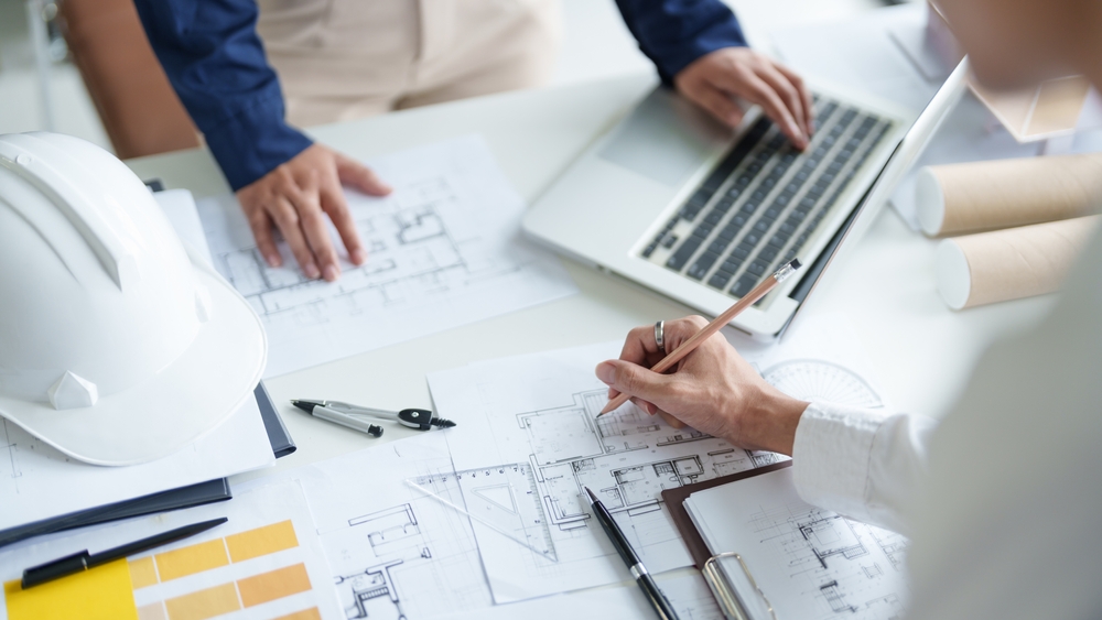 Construction professionals reviewing architectural blueprints at a desk with a laptop, drafting compass, color swatches and a white hard hat.