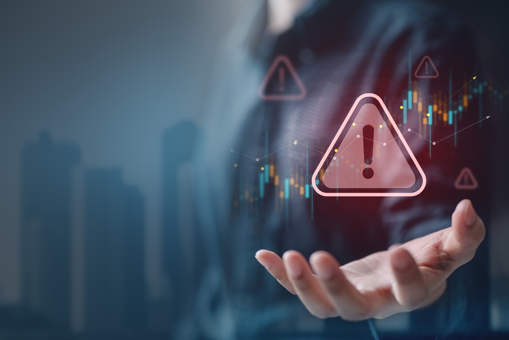 A person holding out a hand with digital warning triangle icons and financial chart data overlaid, symbolizing risk alerts and monitoring, with a blurred city skyline in the background.
