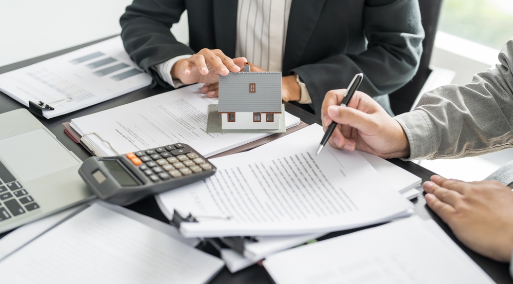Two professionals reviewing and signing contract documents at a desk with a small house model, calculator and laptop, representing a real estate or construction agreement.