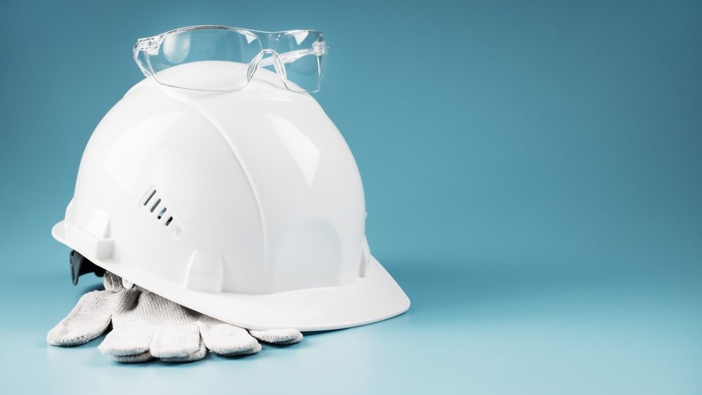 A white hard hat with safety glasses resting on top and work gloves tucked beside it, set against a blue background.