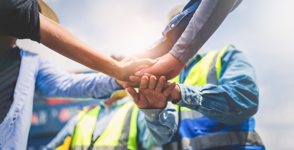 A group of construction workers in high-visibility vests stacking hands together in a teamwork gesture on a jobsite.