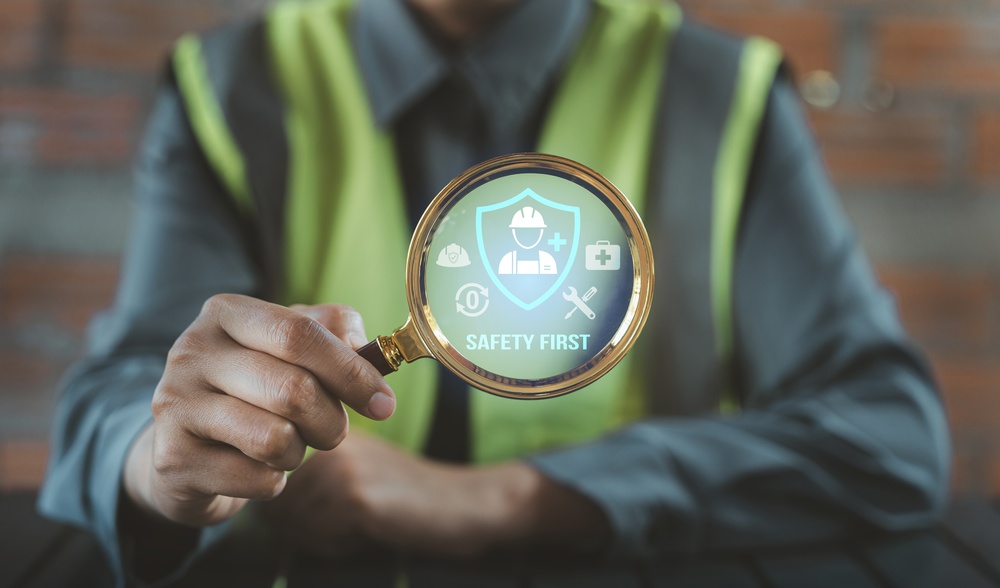 Construction worker in a high-visibility vest holding a magnifying glass over a digital "Safety First" icon featuring a hard hat, first aid kit and shield graphic.
