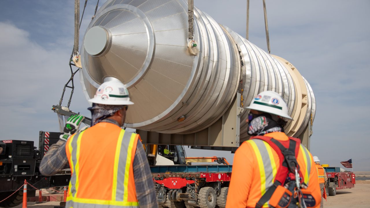 Two workers in hard hats, orange high-visibility vests and face coverings watch as a crane lifts a large insulated cylindrical vessel above a self-propelled modular transporter at a desert construction site.