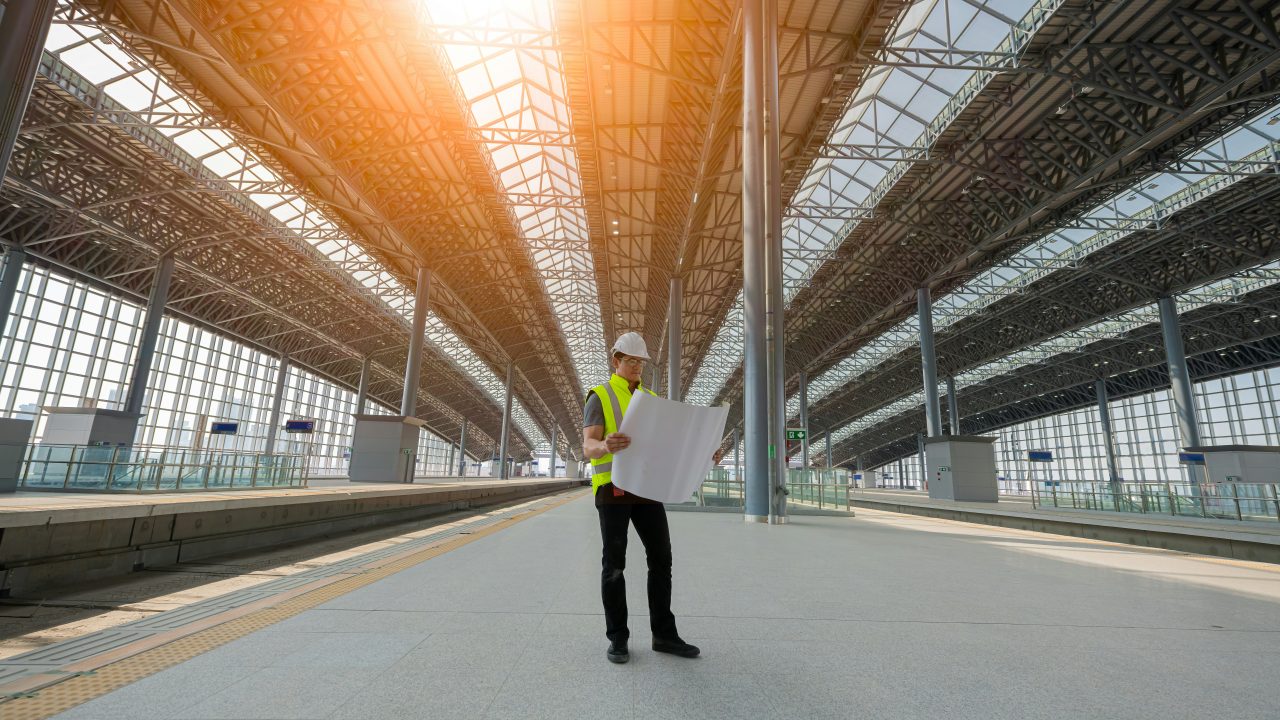 A construction professional in a white hard hat, safety glasses and yellow high-visibility vest reviews large blueprints on an empty platform beneath the steel truss roof and glass canopy of a new train station.