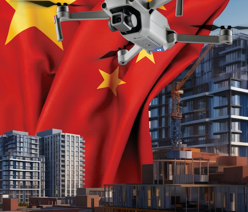 A white quadcopter drone with a camera hovers in front of a large waving Chinese flag, with modern high-rise buildings and a construction crane in the background