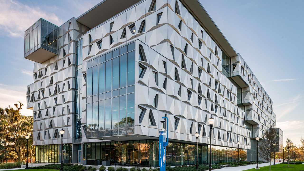 Malachowsky Hall at the University of Florida, a mid-rise building with a faceted metal-panel façade, trapezoidal punched window openings, a large glass curtain wall and a cantilevered glass box, viewed from a lawn with a blue emergency call station in the foreground.
