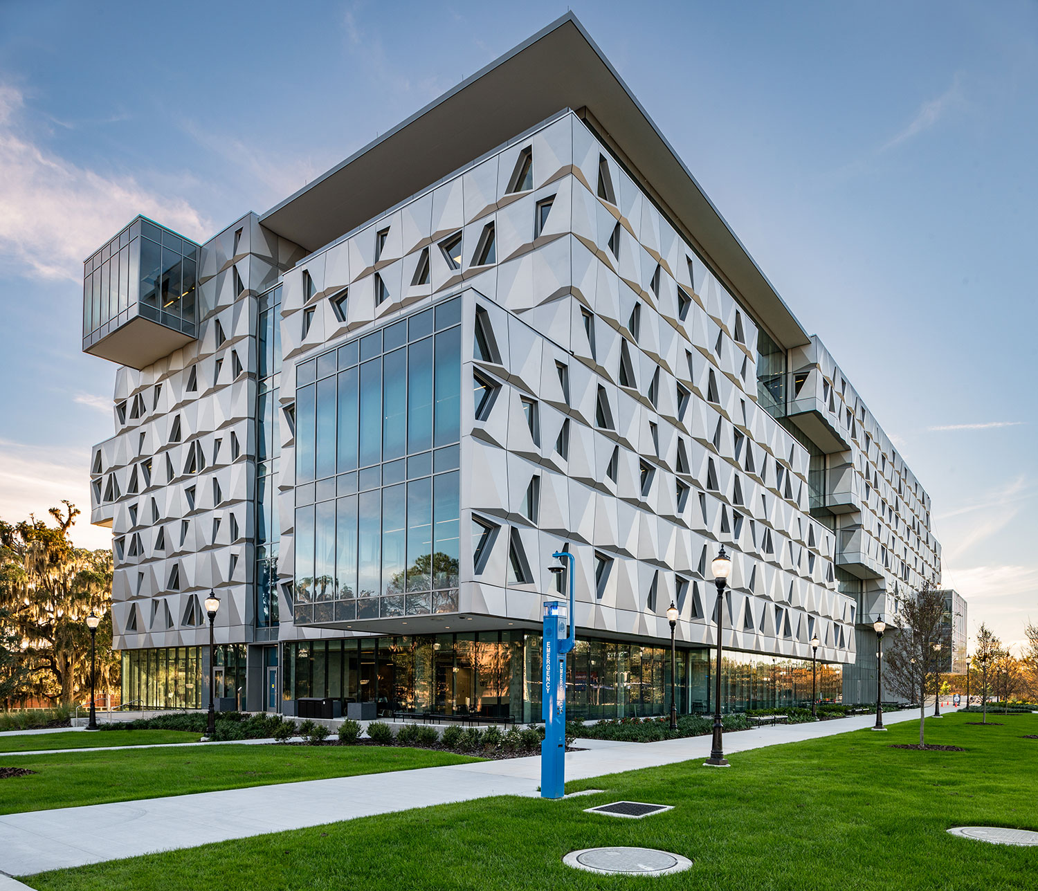 Malachowsky Hall at the University of Florida, a mid-rise building with a faceted metal-panel façade, trapezoidal punched window openings, a large glass curtain wall and a cantilevered glass box, viewed from a lawn with a blue emergency call station in the foreground.