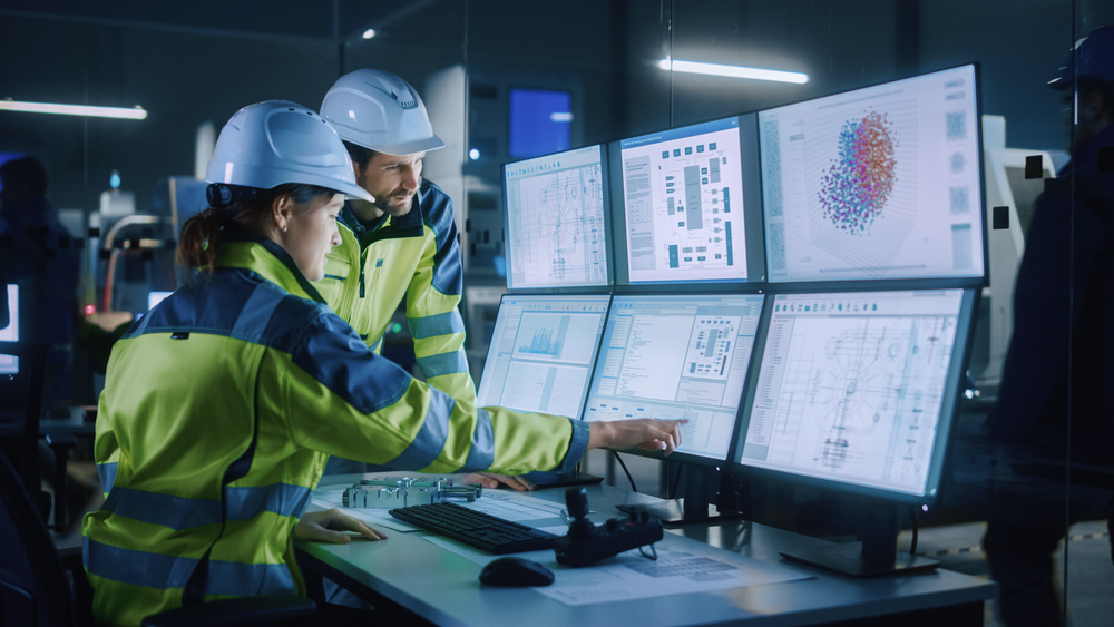 Two workers in hard hats and high-visibility jackets review technical drawings and data on a bank of computer monitors in a control room, with one pointing at a screen.