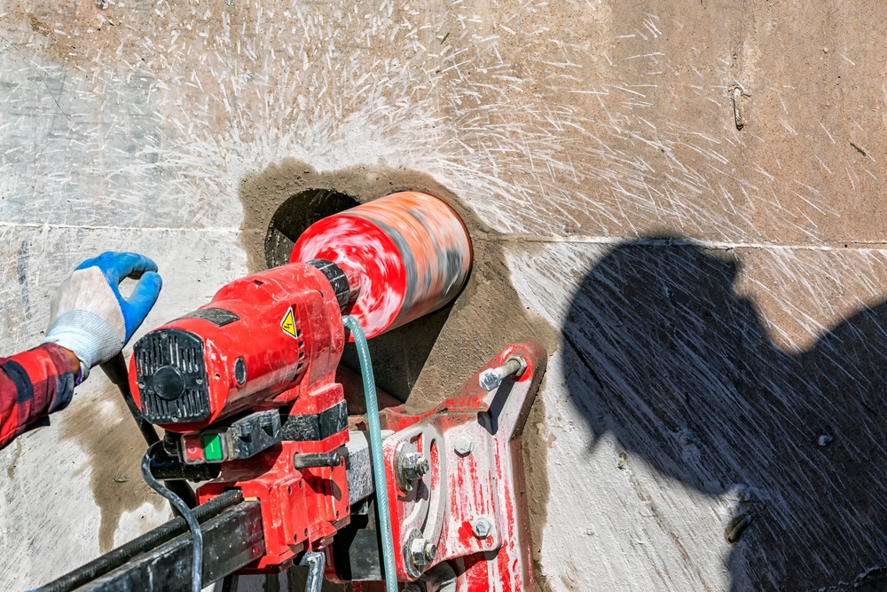 A red core drill rig mounted to a concrete wall, drilling a large hole as water sprays outward, with a gloved hand steadying the equipment.