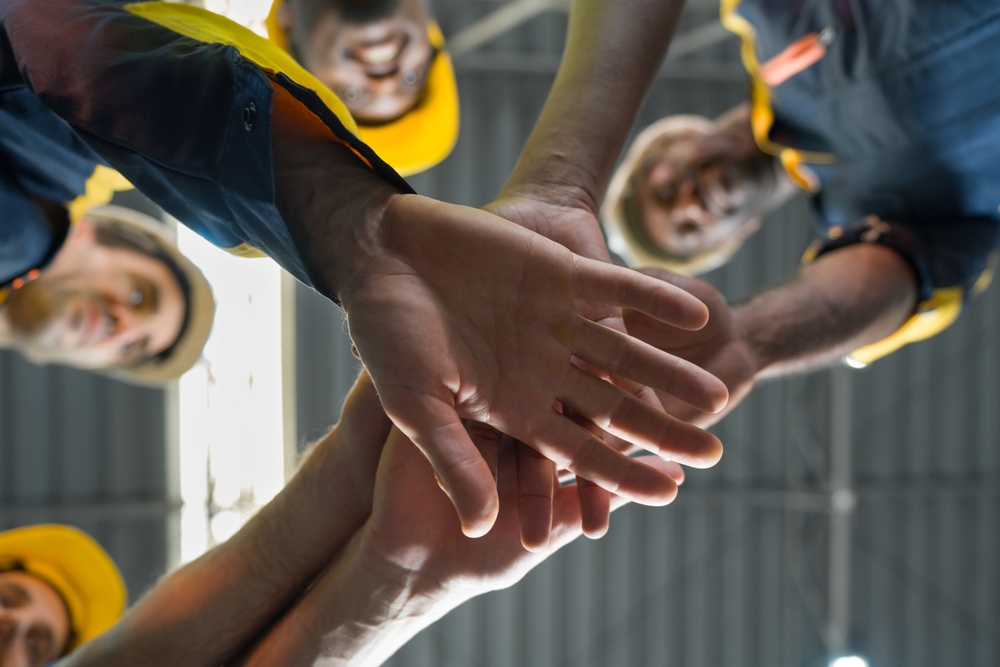A low-angle view of construction workers in yellow hard hats smiling down as they stack their hands together in a team huddle inside an industrial building.