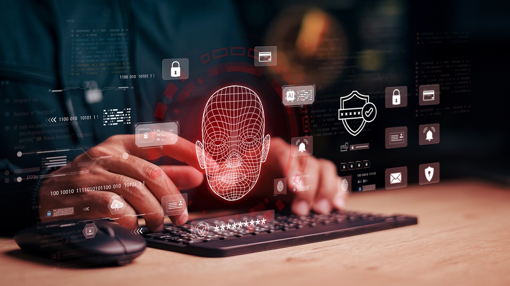 A person types on a keyboard beside a mouse while a glowing red wireframe face floats above the keys, surrounded by digital icons for padlocks, a shield with a checkmark, a password field, an AI chip, a bell, a credit card, an envelope and lines of binary code.