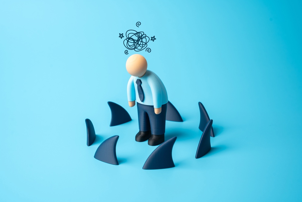A small figure of a stressed man in a shirt and tie with a scribbled cloud above his head, surrounded by dark shark fins on a bright blue background.