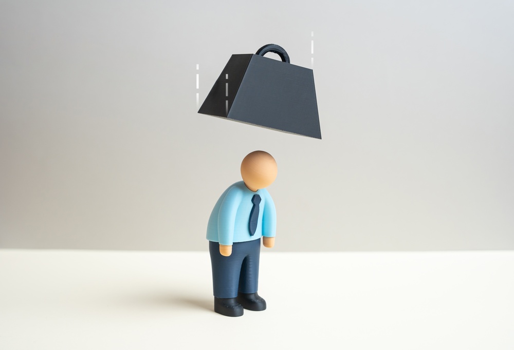 A small figurine of a man in a light blue shirt, tie and dark pants stands with his head bowed while a heavy black weight hovers above him, symbolizing stress or burden.