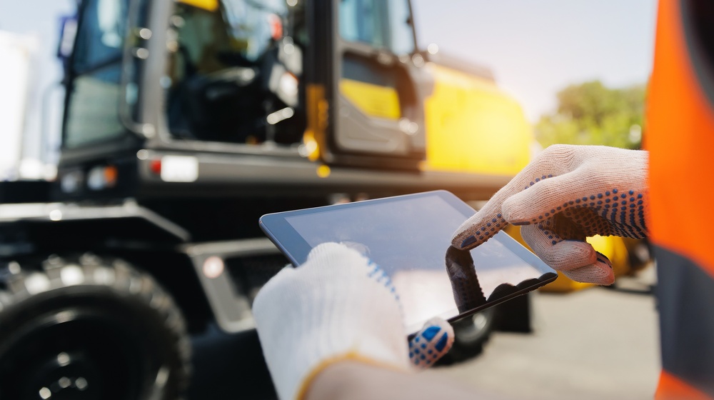 “Close-up of a worker in an orange high-visibility vest and dotted work gloves using a tablet in front of a yellow wheeled excavator.