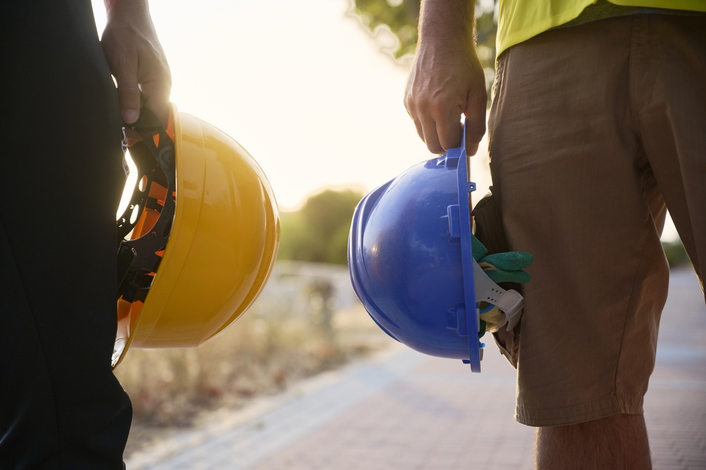 Close-up of two construction workers standing side by side outdoors at sunset, one holding a yellow hard hat and the other a blue hard hat at their sides, with green work gloves tucked into a pocket.