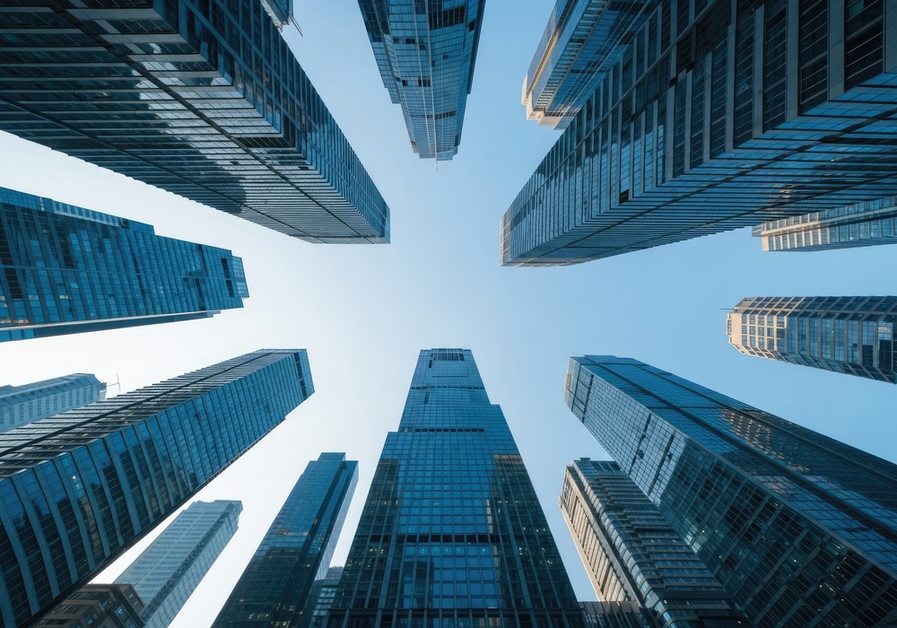 Low-angle view looking up at a cluster of modern glass skyscrapers converging toward a clear blue sky.