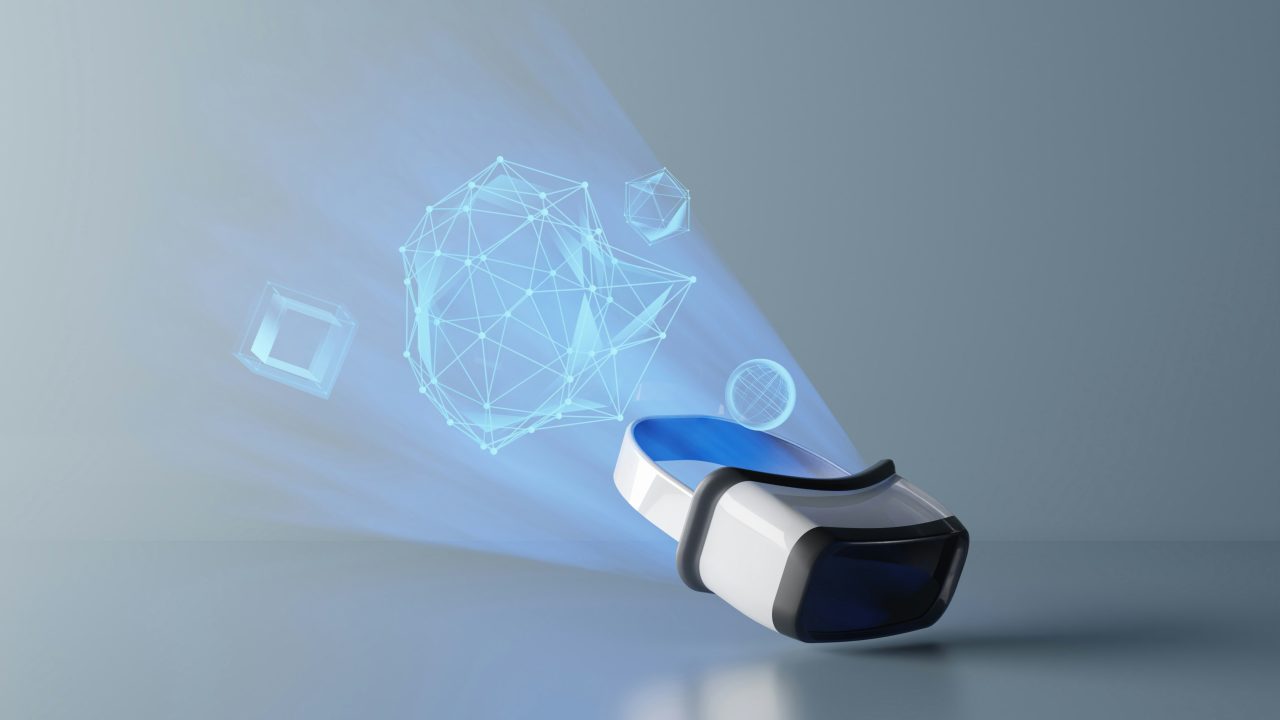 A white virtual reality headset resting on a reflective surface, projecting a beam of blue light that contains glowing wireframe shapes, including a large polyhedral mesh, a cube and a sphere.